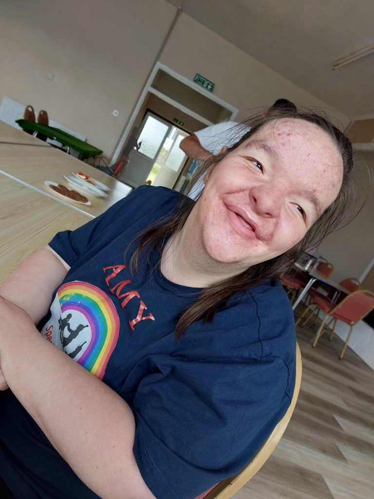 Girl wearing rainbow tee-shirt is sat at a table and smiles at camera 