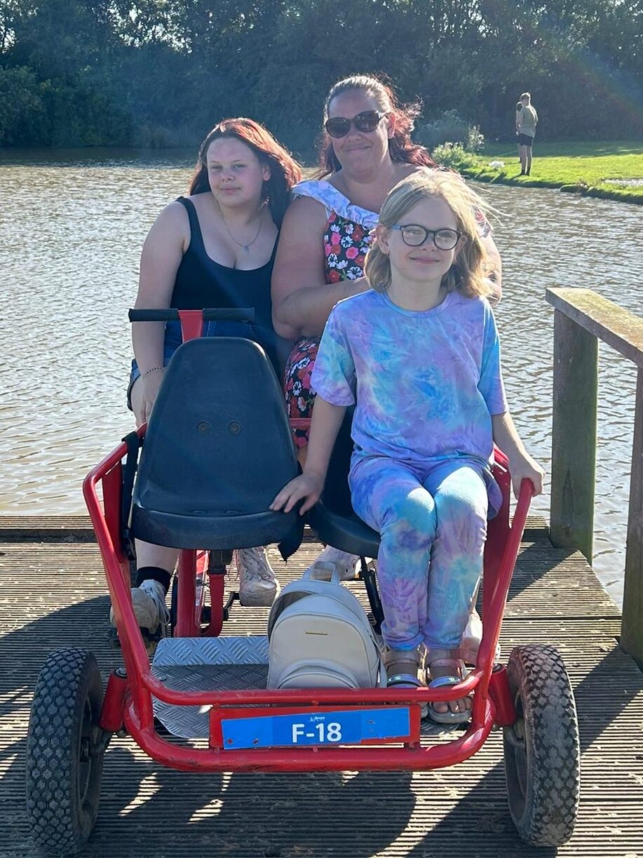 Lucy, Molly and Sam are on a four-person bike, behind them is a lake. It is sunny and all three are sat on the bike, smiling.