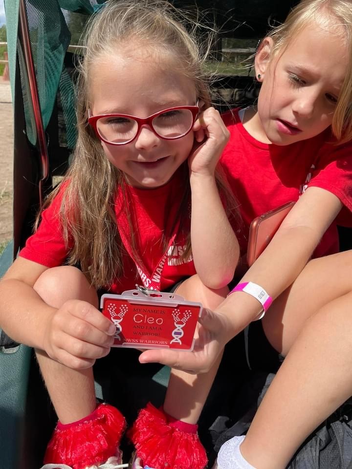 Young girl wearing glasses sits next to her sister and holds a badge with her name on up to the camera.