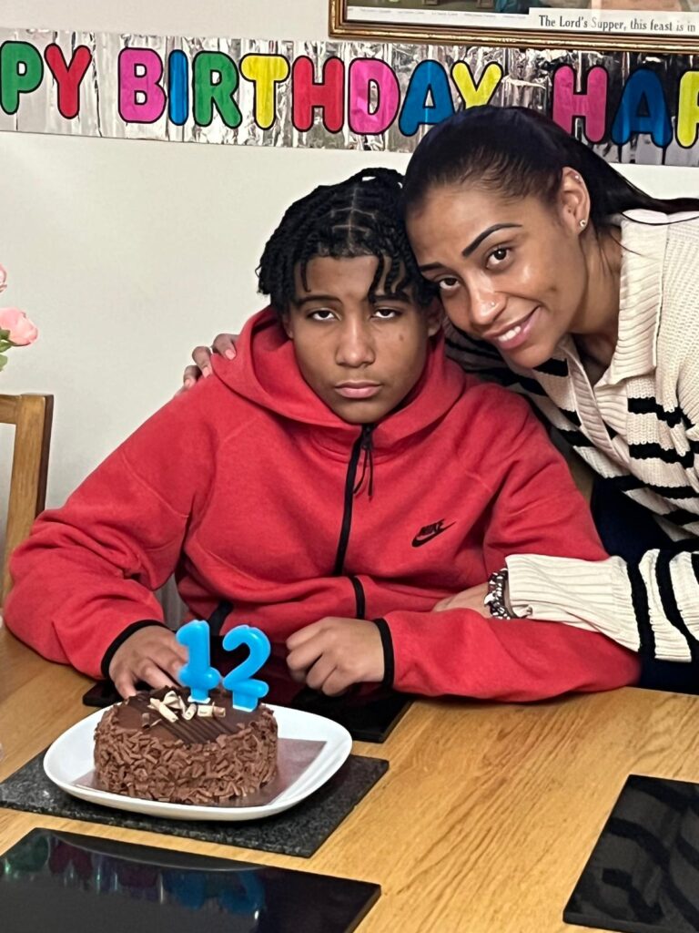 Ryanne and Derion are next to a dining table. There is a 'Happy Birthday' banner in the background, and a chocolate cake in front of them with candles reading 12. Ryanne is smiling with her arm around Derion, who is wearing a red zip-up hoodie.