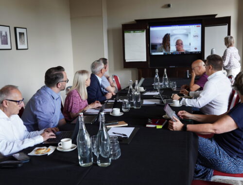 A group of people sitting in front of laptops around a conference table. Two people are visible on a screen, Zoomed into the meeting.