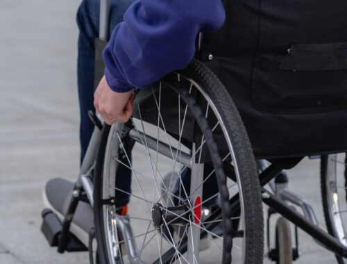 The back of a wheelchair with the wheelchair user's hand visible on the wheel.