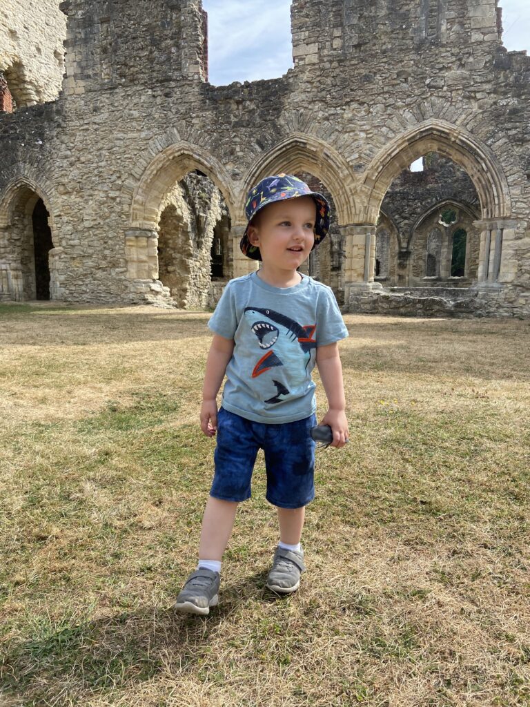 Little boy wearing shorts, tee shirt and hat stands on grass outside an old ruin on a sunny day