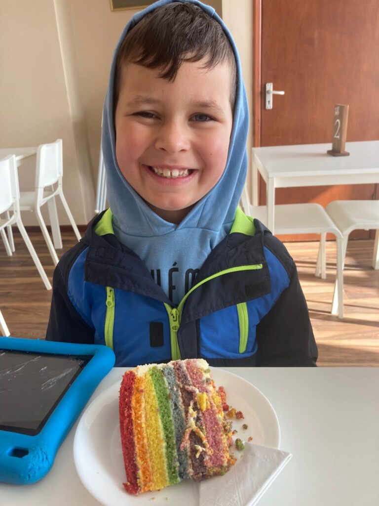 Cody is sat in a cafe with a slice of rainbow cake in front of him. He is smiling widely at the camera, wearing a hood. He has dark brown hair.