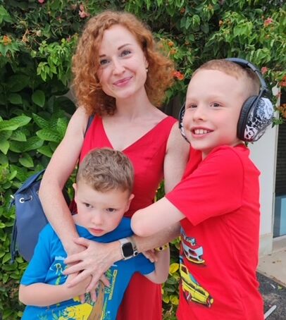 Irina, David and Eli are on holiday. Irina is wearing a red dress, with her arms around Eli. David linked his arm into Irina's, and wears headphones.