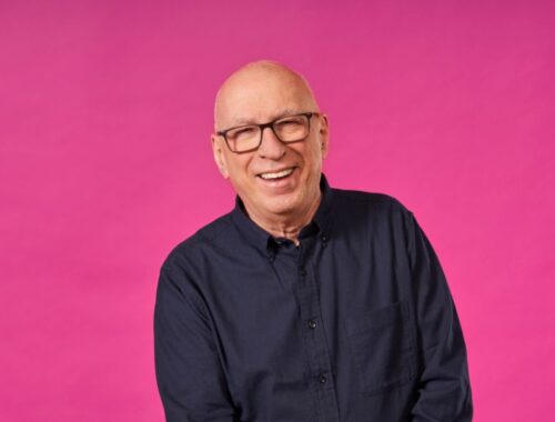 A man with glasses who is smiling stood against a pink background