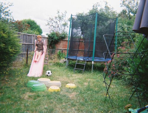 A little boy at the top of a small slide in a garden. Behind him is a trampoline