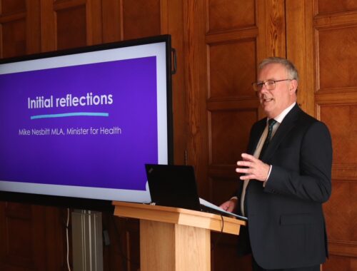 Minister Mike Nesbitt speaking in front of a screen that says 'Initial reflections, Mike Nesbitt MLA, Minister for Health.'