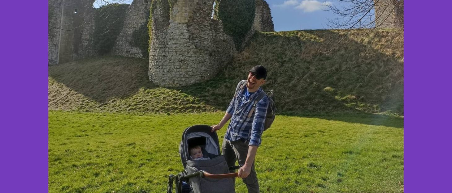 A photo of Nathan and his daughter in the pram smiling for the camera in the park.