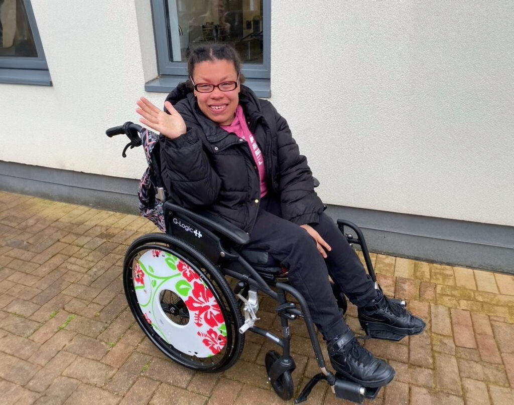 Nur in a wheelchair smiling and waving at the camera