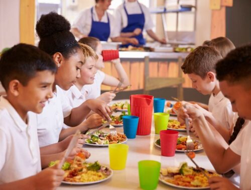 Children in a primary school sat at a school lunch table with kitchen team behind them.