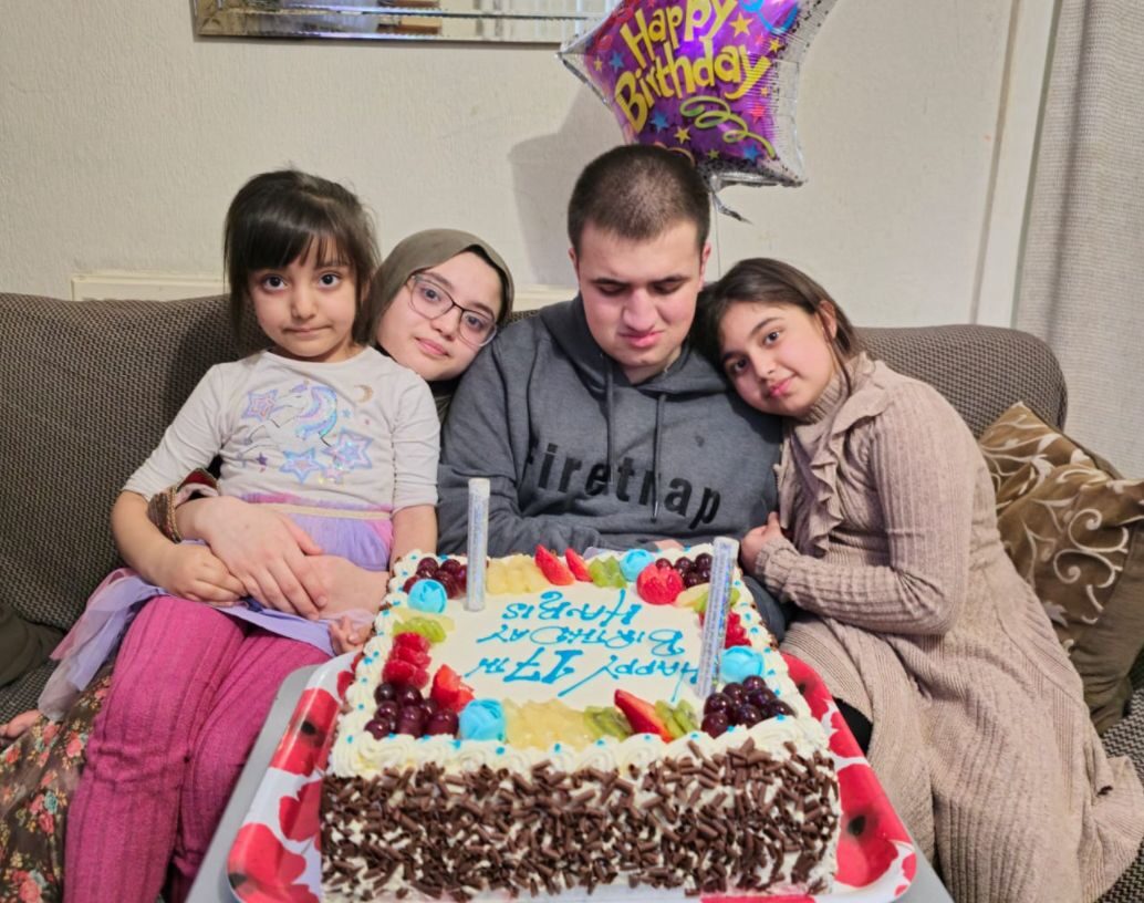 A group of four children sitting around a birthday cake with a birthday balloon in the background.
