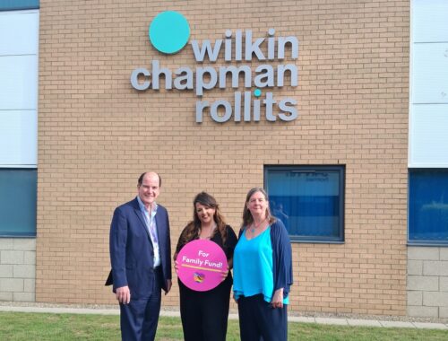 Three people standing outside in front of a sign that says 'Wilkin Chapman Rollits.' The person in the middle is holding a pink sign that says 'For Family Fund!', with a heart below it.