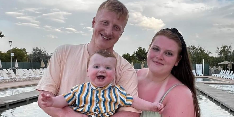 Matthew is holding Theodore, who is holding his arms out and laughing joyously. Sophie is also holding Theodore with Matthew. They are on holiday, abroad.