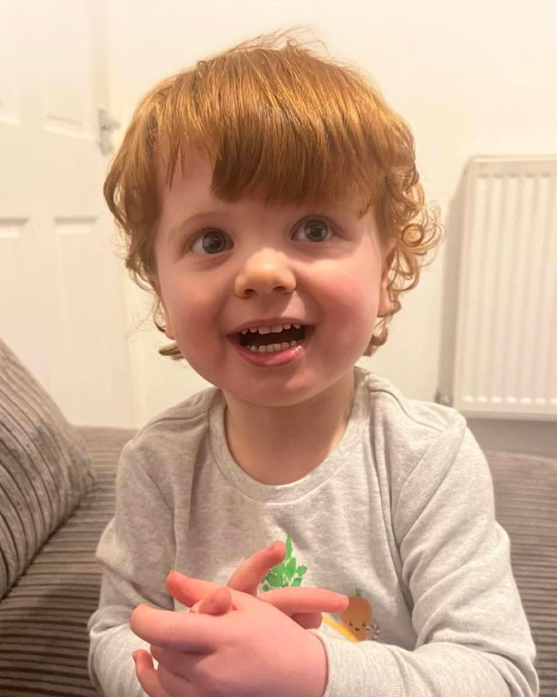 Theodore is holding his hands together and smiling. He has curly, ginger hair and is looking to the side of the camera.