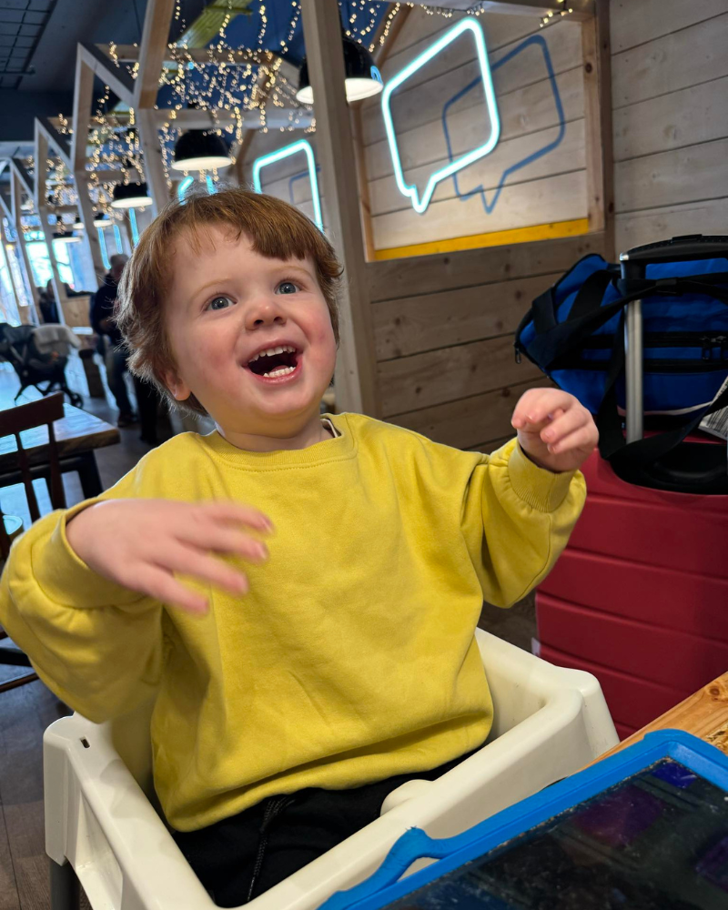 Theodore is sat at a table at a restaurant. He is holding his hands in the air and giving an open-mouth smile.