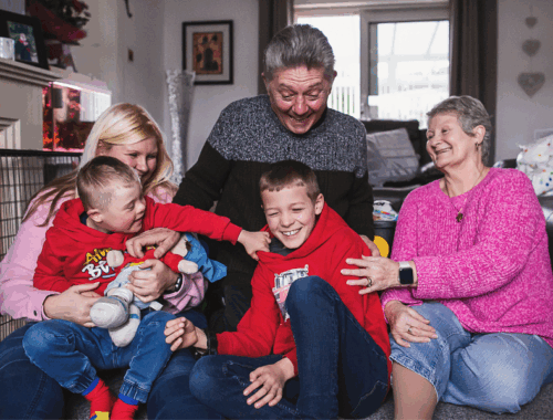 A family is sat on the floor in the living room. The grandfather, grandmother and mother and next to them two young boys. The youngest is pulling the his brother's hoodie. Everyone is laughing.