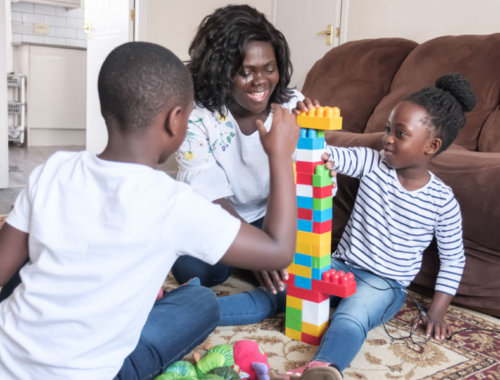 A photo of a woman sat on the floor next to her son and daughter. They are building a tower using lego blocks.
