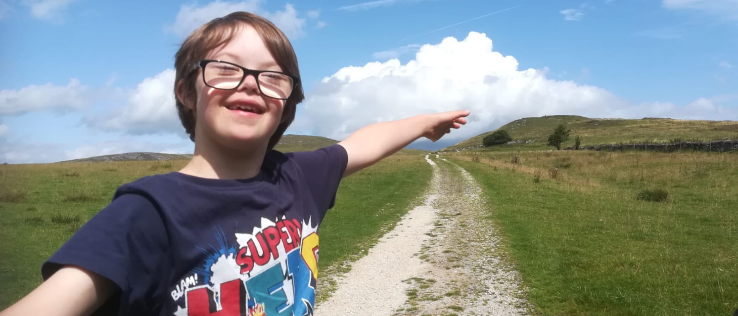 Alex is outside on a long walking track. He is surrounded by green fields and has his arms spread widely. He wears a t-shirt that says 'super hero', has glasses and floppy brown hair. He is smiling.