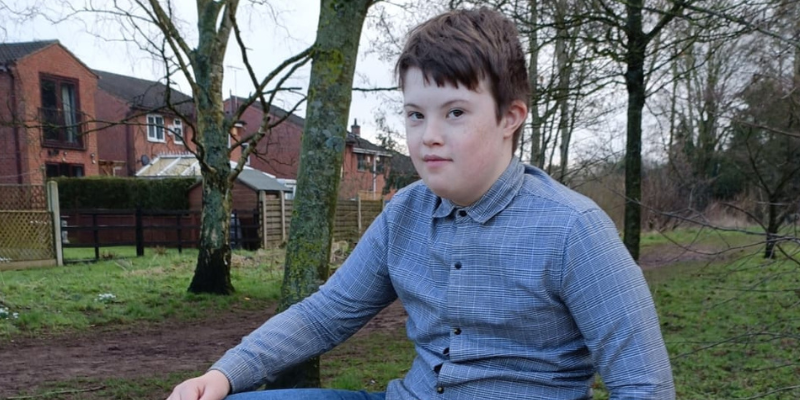 Alex wears a smart shirt and he is pictured in a park. He has short brown hair and is looking directly at the camera.