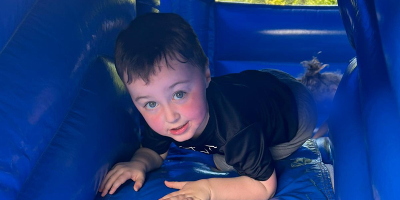 Arthur, who has bright blue eyes and short brown hair, is climbing through a blue inflatable and is peering up towards the camera.