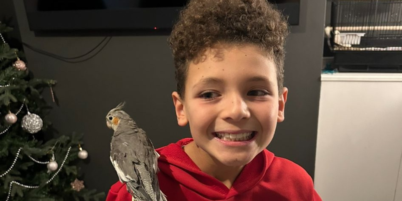 Blake is wearing a red hoodie, with curly hair and there is a parrot sat on his shoulder. He is smiling a wide smile, looking to his right at the parrot.