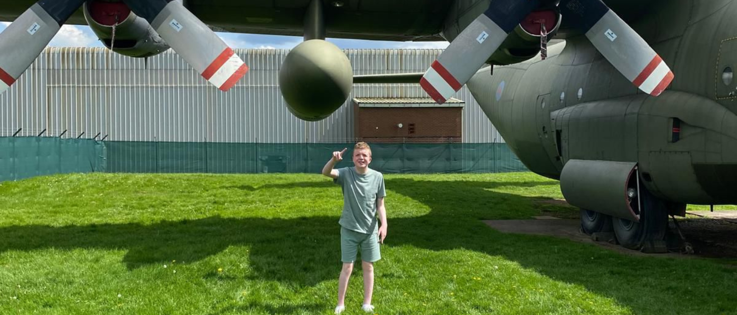 Lennon is stood beneath a large RAF plane on a sunny day. He is pointing towards the camera.