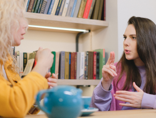 Two women sitting at a table in front of a book shelf, using their hands to sign to each other