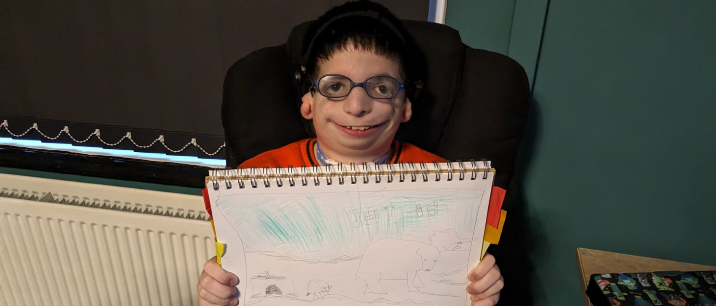 Cameron is smiling proudly, holding up his drawing. He is wearing glasses, a red t-short and sits on an office chair at home.