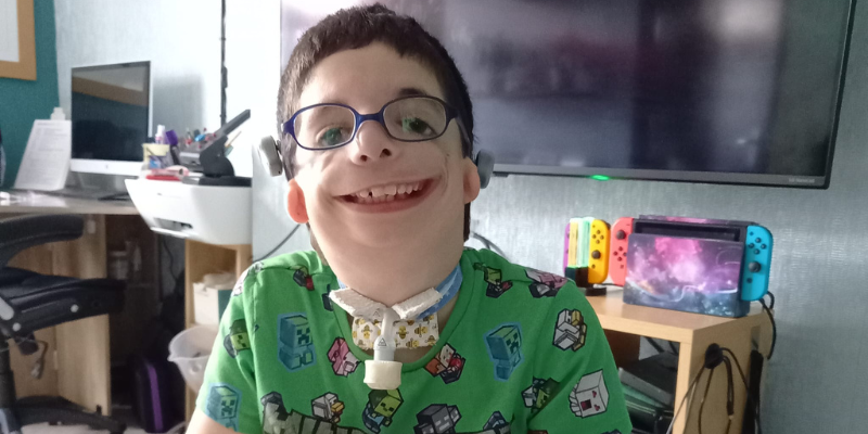 Cameron is smiling widely. He wears glasses and a mine craft t-shirt, and you can see his tracheostomy. He has short dark hair.