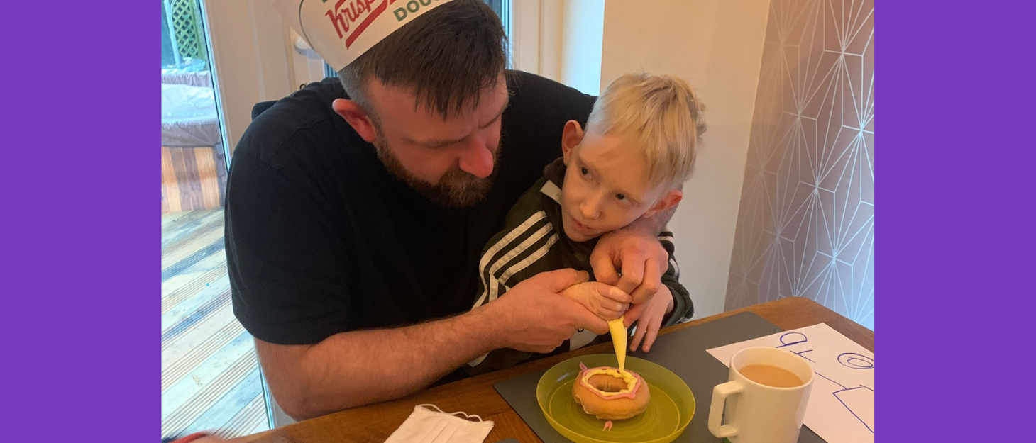 Kris is sat at the dinner table with Mason on his lap. He is wearing what looks like a party hat, and he is decorating a doughnut with Mason.
