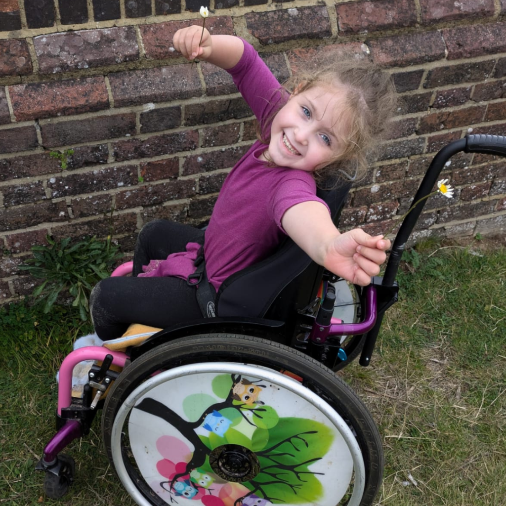 Charlotte is sat in her wheelchair holding daisies in each hand and smiling.