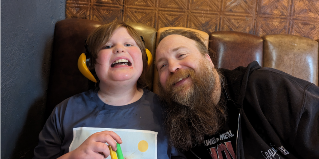 Connor is sat at a table wearing headphones and holding felt-tips, next to his dad Kris. Kris is leaning into him and they are both smiling.