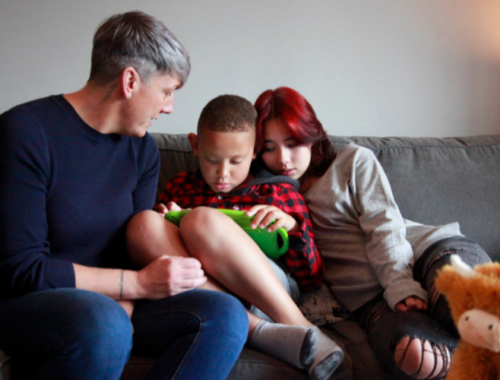 A woman is sat on the sofa next to a boy and a girl. The boy is holding a tablet and is looking at the screen.