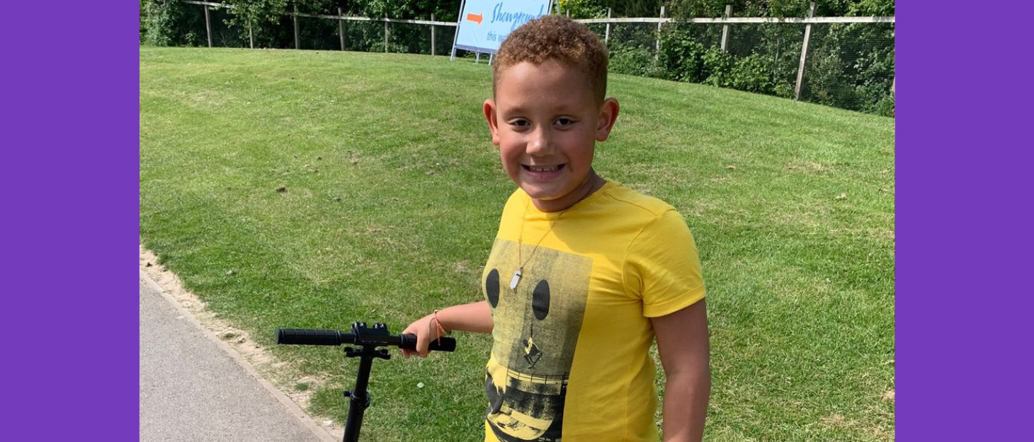 Boy wearing yellow teeshirt stands on path holding scooter and smiling at the camera