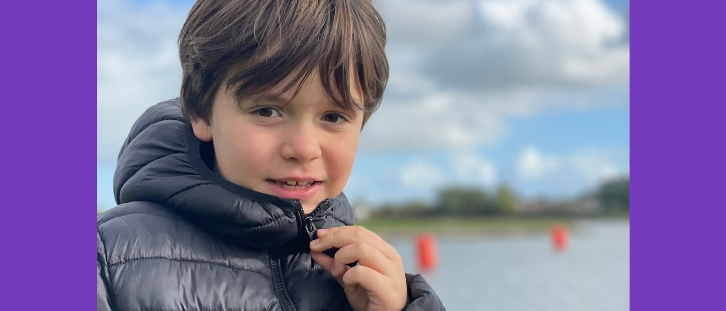 Young boy zips up his black coat in front of lake on cold day.