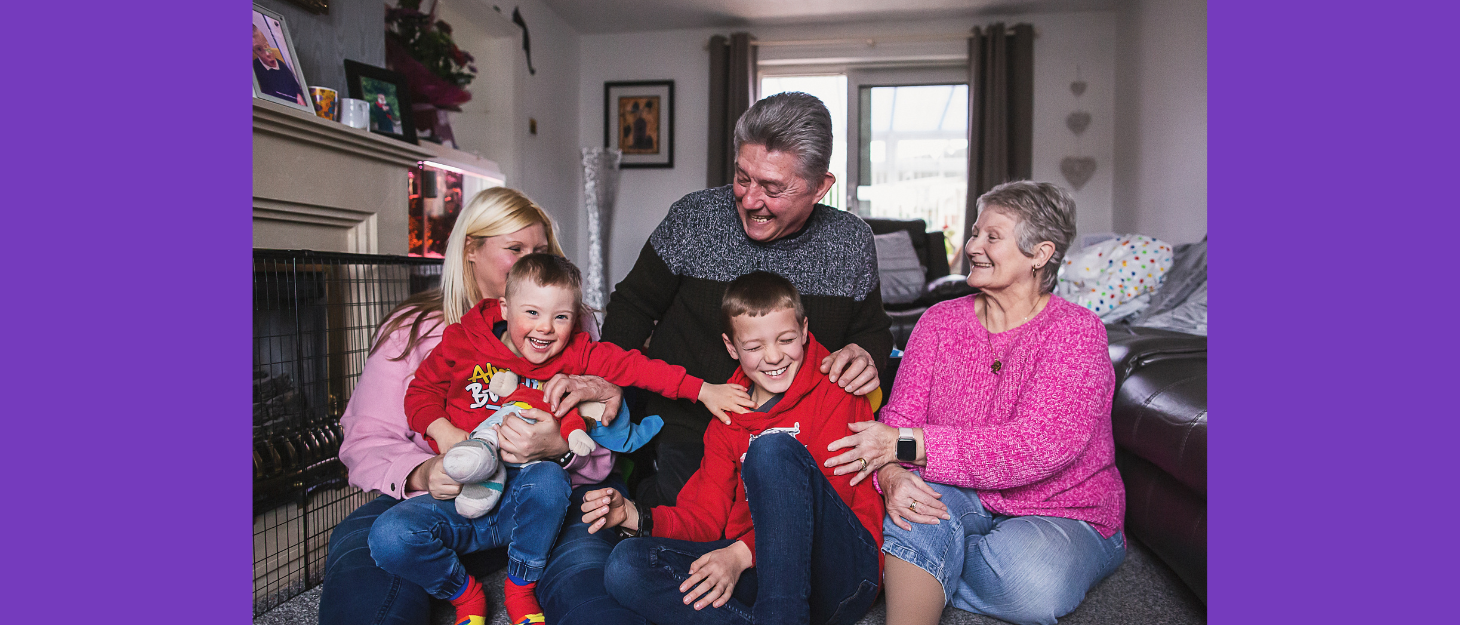 Family with two boys, mum and grandparents sit on the floor of the living room, tickling each other and laughing.