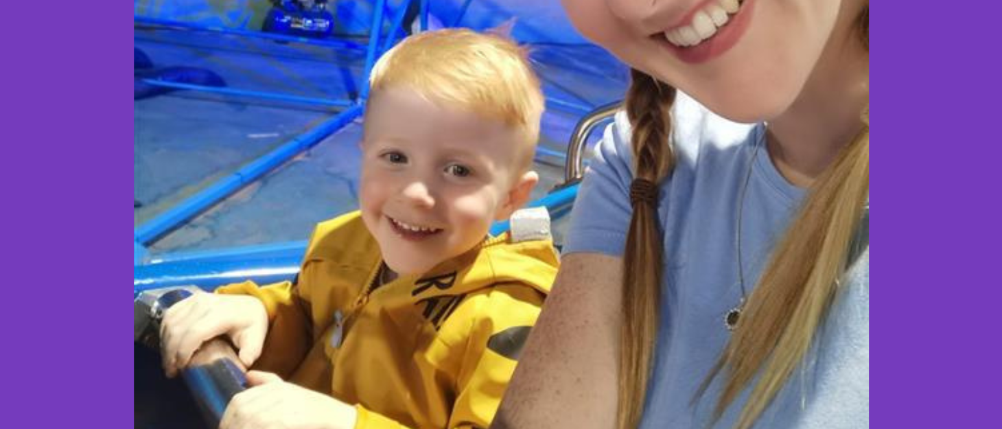 Young boy with blonde hair and wearing yellow jumper sits next to his mum on ride and smiles for a selfie