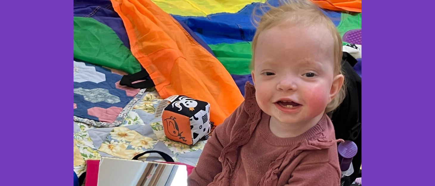 Baby girl sitting amongst sensory toys, smiles at camera