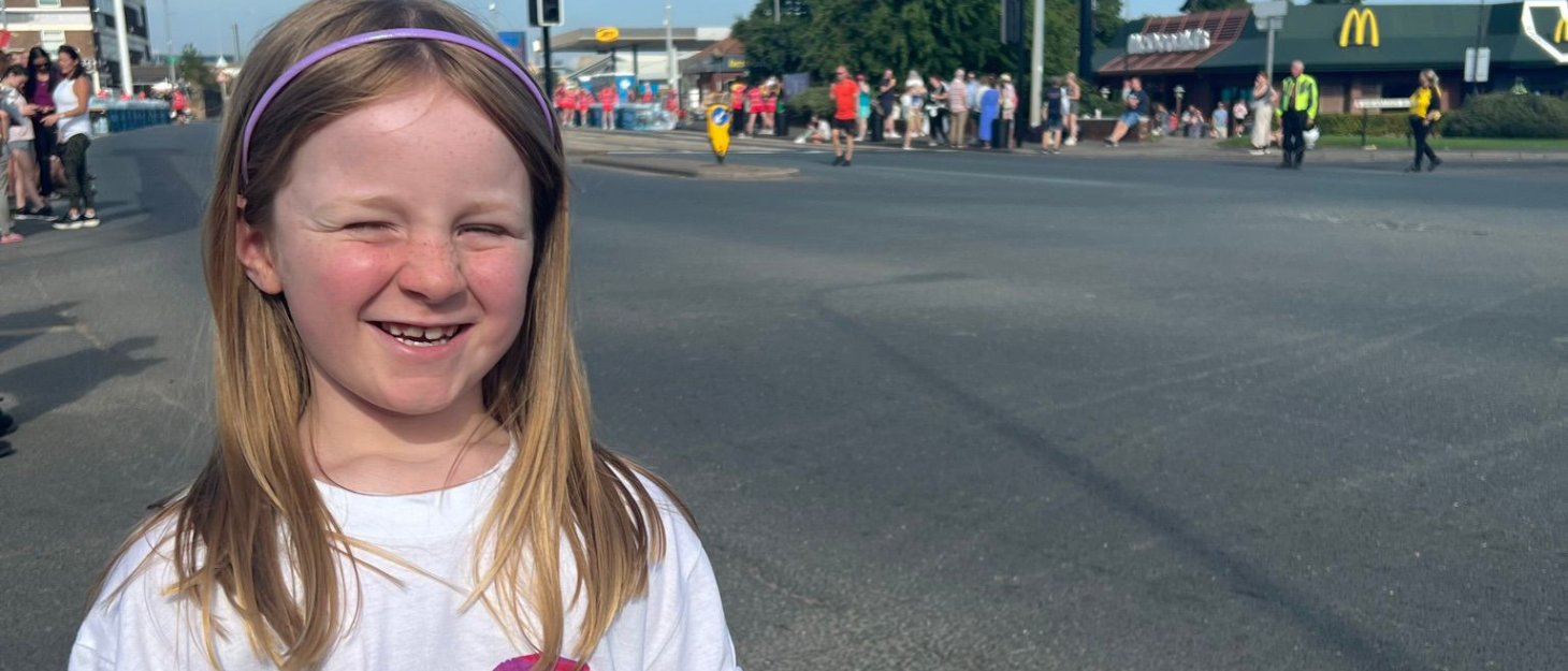 Daisy is stood outside on a sunny day, so she is squinting slightly whilst smiling. She is wearing her Family Fund t-shirt, and is waiting for Great North Runners to come past.