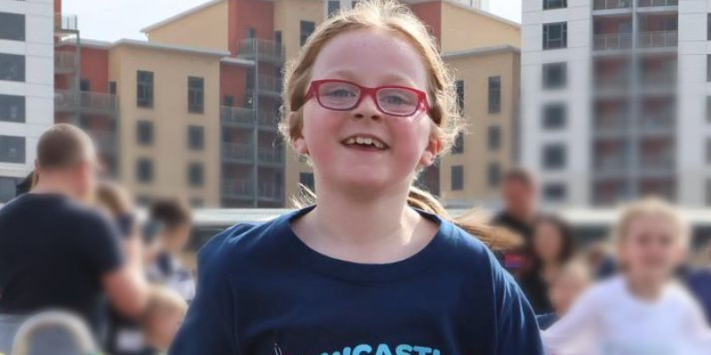Daisy has her hair tied back, as she is running with all her might at the Mini Great North Run. She is surrounded by other runners and families, and is smiling at the camera.