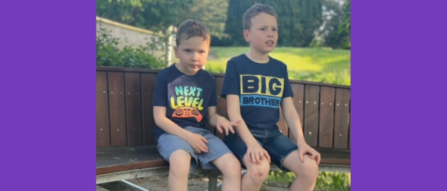 Eli and David are sat on a bench outdoors. It is summer time and they are sat looking into the distance, wearing shorts, trainers and t-shirts.