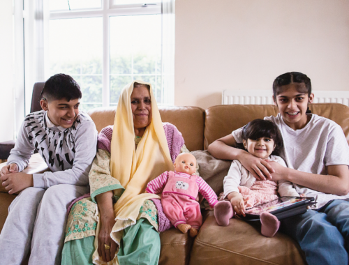 A woman is sat on the sofa next to her children: a teenage boy, a teenage girls and a young girl holding an iPad. They are smiling.