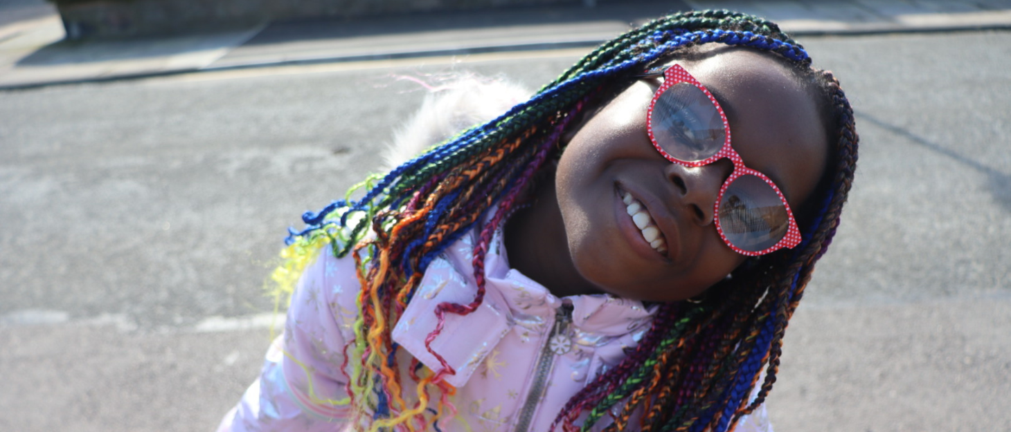Elizabeth is smiling with her head tilted to the side outside. She is wearing a pink coat, red polka-dot glasses and rainbow-coloured braids.