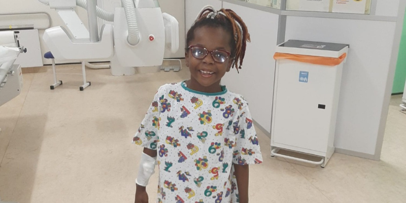 Elizabeth is smiling, stood in hospital with a bandage on her arm. She wears a hospital gown with illustrations and numbers on it.