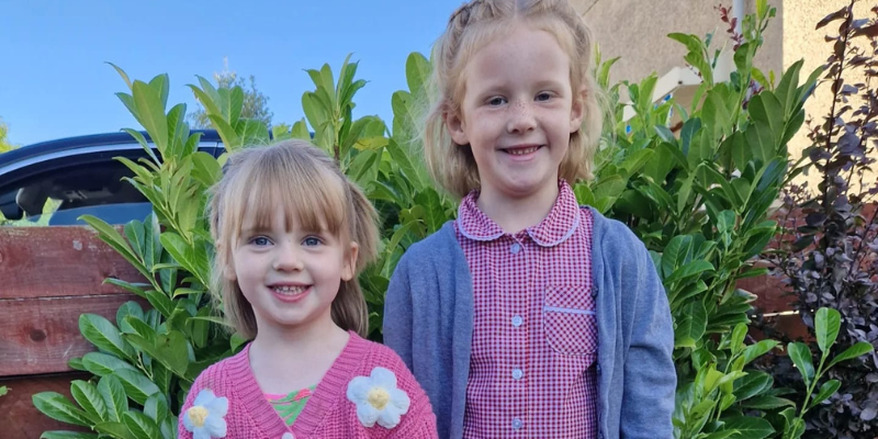 Eve and Olivia are stood in front of greenery and they are both smiling. Both girls have blond hair, smile at the camera, and are dressed with summer clothes.