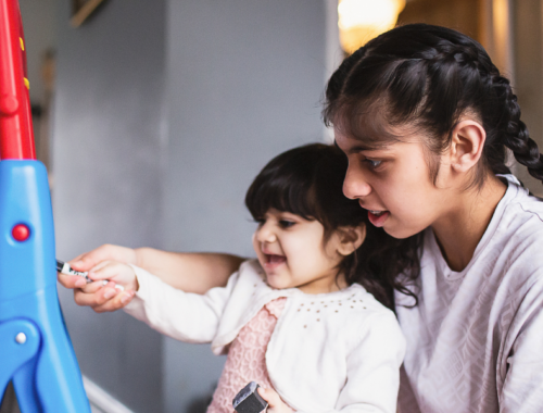 Two siblings writing on a blackboard, the older helping the little one