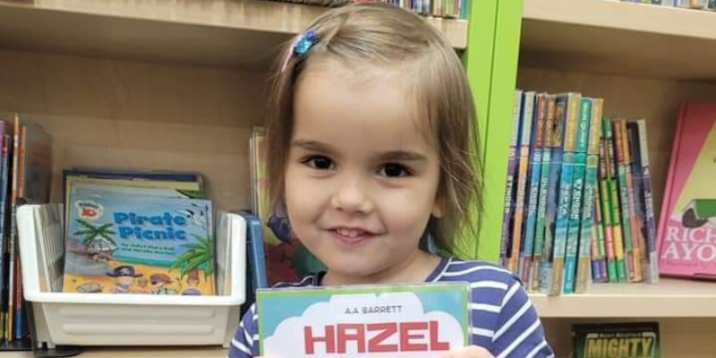 Hazel is holding a book that reads 'Hazel' and is stood in front of a bookcase, smiling.