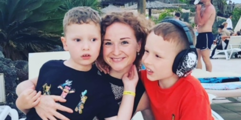 Irina, David and Eli are on holiday. Eli is sat on Irina's knee, holding her face, and David is sat closely beside her. Irina is smiling.