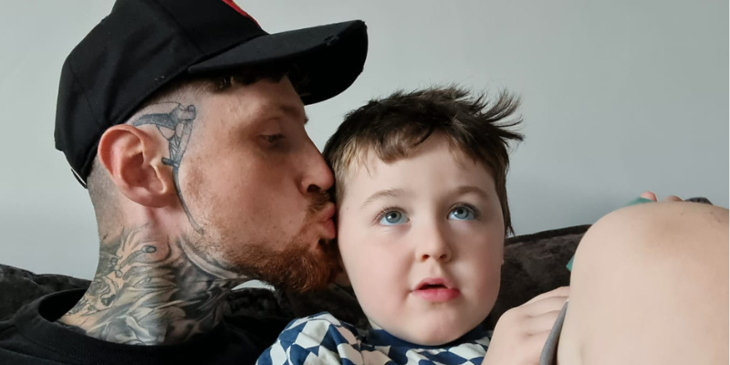 Joe and Arthur are sat on the couch. Arthur is looking up, and Joe is kissing Arthur's temple. Arthur has short brown hair and blue eyes, and Joe has his eyes closed, with tattoos on his face and neck, and a black cap.